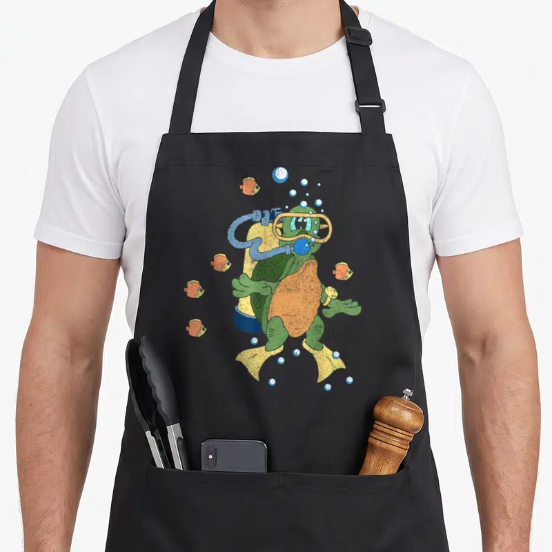 Tablier de cuisine noir avec une tortue verte équipée d'un scaphandre et de palmes jaunes, entourée de poissons orange et de bulles.
