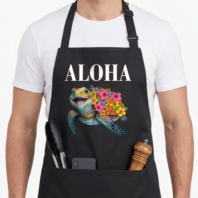 Tablier de cuisine noir avec une tortue souriante ornée de fleurs Aloha et le mot Aloha en lettres blanches.