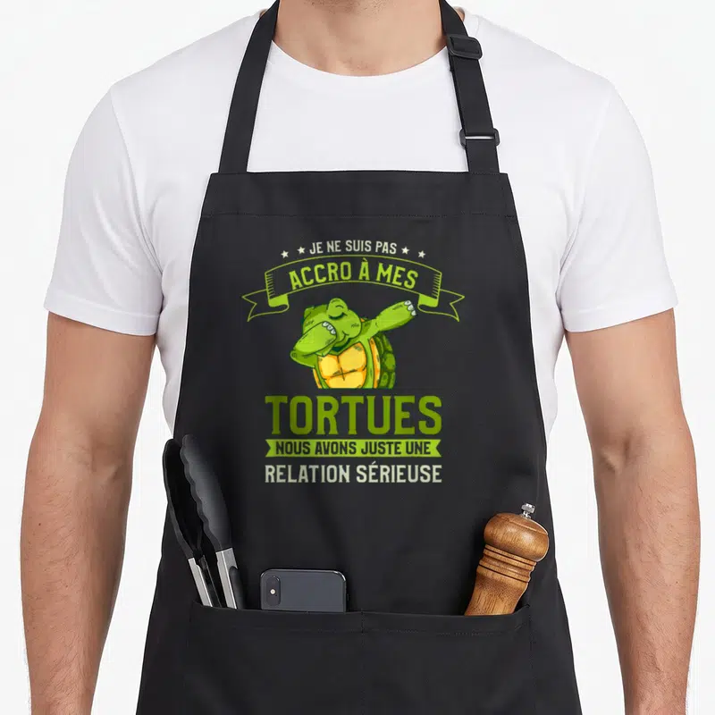 Tablier de cuisine noir avec une tortue verte stylisée effectuant un mouvement dab et du texte humoristique imprimé en vert et blanc.