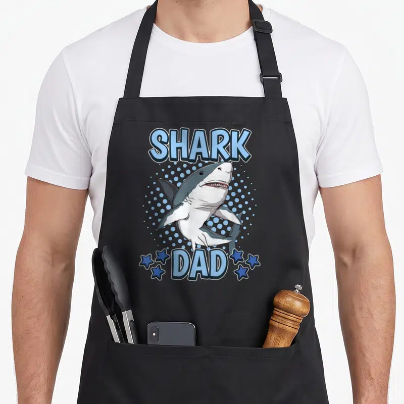 Tablier de cuisine noir avec illustration d'un requin souriant et le texte Shark Dad en bleu.