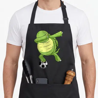 Tablier de cuisine noir avec une tortue verte en train de faire un dab sur un ballon de football noir et blanc.