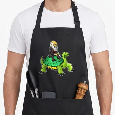 Tablier de cuisine noir avec illustration colorée de Charles Darwin en costume montant sur le dos d'une grande tortue verte des Galapagos.