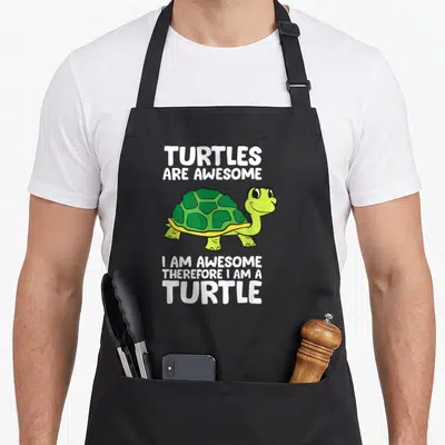 Tablier de cuisine noir avec une tortue verte souriante et un texte humoristique en anglais sur le devant.