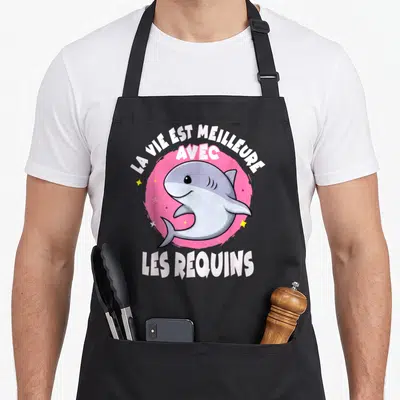 Tablier de cuisine noir avec un requin cartoon gris sur fond rose et texte blanc La vie est meilleure avec les requins