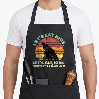 Tablier de cuisine noir avec aile de requin sur fond de soleil rayé et texte humoristique en anglais.