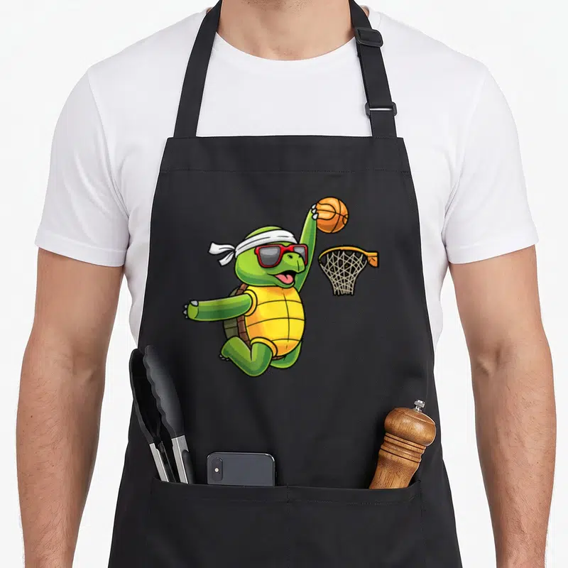 Tablier de cuisine noir avec une tortue verte sautant vers un panier de basket avec un ballon orange.