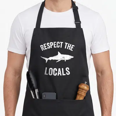 Tablier de cuisine noir avec illustration de requin blanc et texte Respect The Locals en typographie usée.