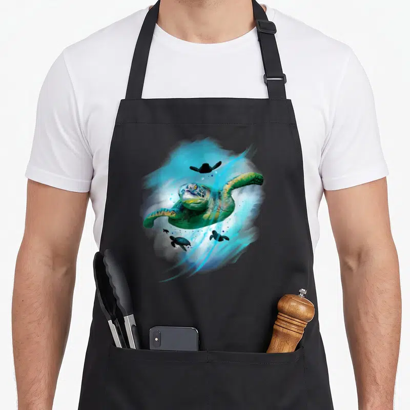 Tablier de cuisine noir avec une tortue de mer peinte dans des tons bleus et verts, entourée de bébés tortues.