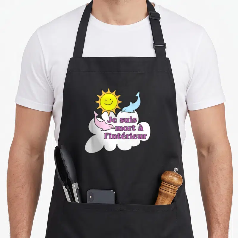 Tablier de cuisine noir avec deux dauphins rose et bleu, un soleil jaune et une bulle de dialogue blanche contenant du texte violet.