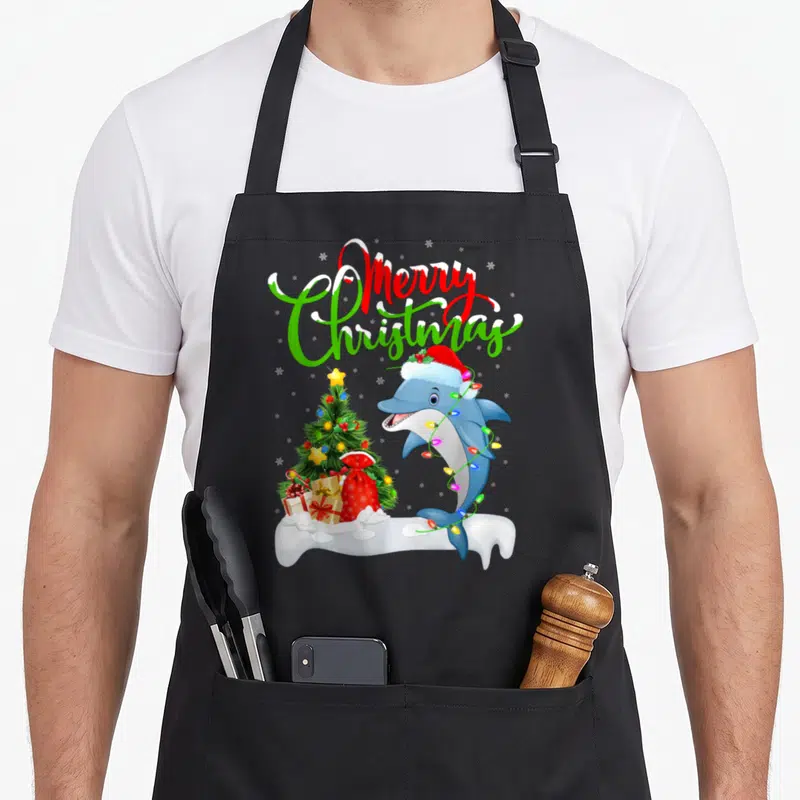 Tablier de cuisine noir avec un dauphin festif portant un bonnet de Père Noël et des guirlandes lumineuses près d'un petit sapin de Noël.