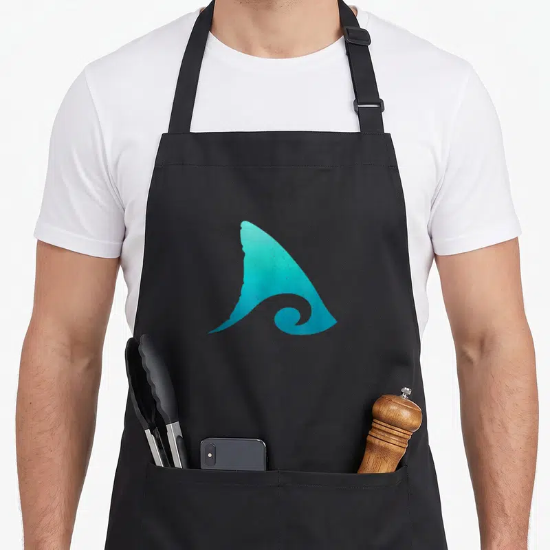 Tablier de cuisine noir avec une illustration stylisée d'une aile de requin teck émergeant d'une vague.