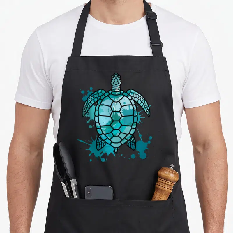 Tablier de cuisine noir avec une illustration de tortue de mer turquoise et des éclaboussures artistiques.