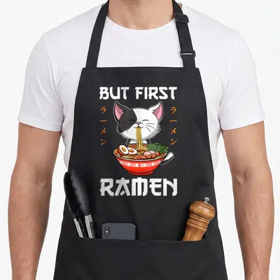 Tablier de cuisine noir avec un chat bicolore mangeant du ramen et le texte But First Ramen.