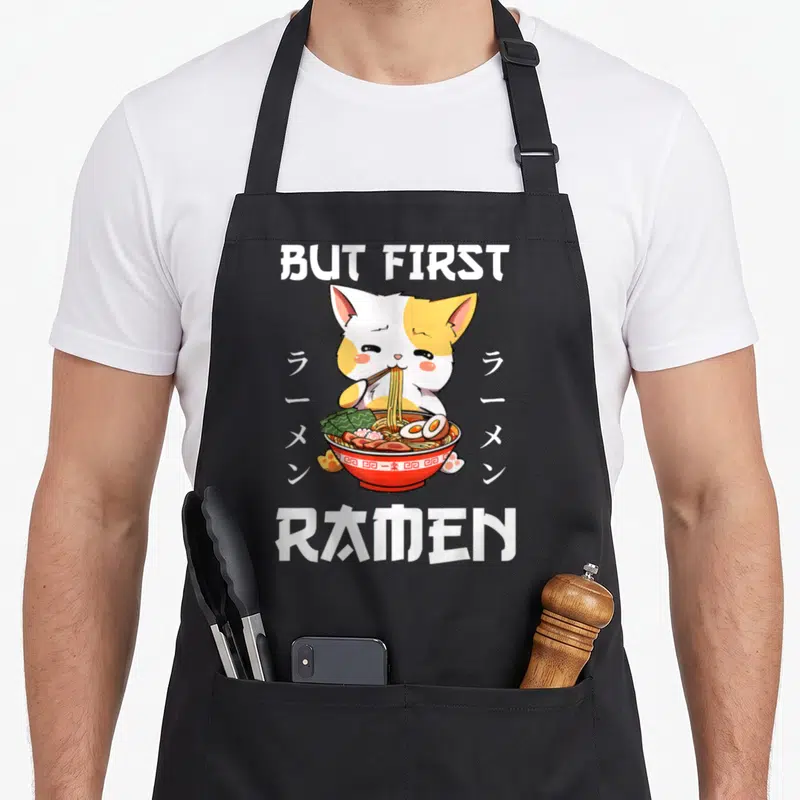 Tablier de cuisine noir avec illustration d'un chat blanc et orange dévorant un bol de ramen rouge.