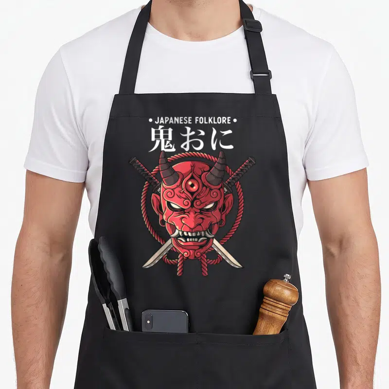 Tablier de cuisine noir avec masque Oni rouge, cornes, épées croisées et texte japonais.