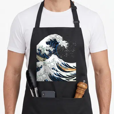Tablier de cuisine noir avec une reproduction de La grande vague au large de Kanagawa par Hokusai, montrant les bateaux et l'écume.