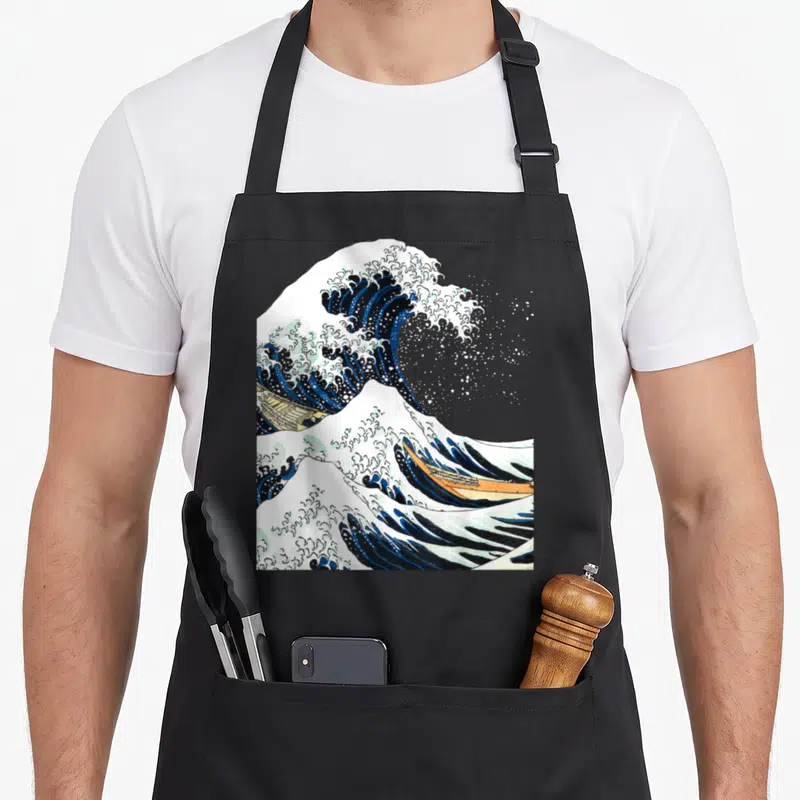 Tablier de cuisine noir avec une reproduction de La grande vague au large de Kanagawa par Hokusai, montrant les bateaux et l'écume.