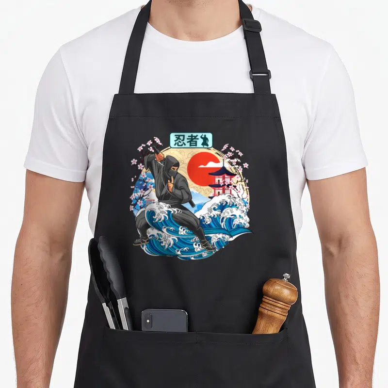 Tablier de cuisine noir avec illustration ninja sur vague bleue, soleil rouge et fleurs de sakura dans un style japonais vintage.