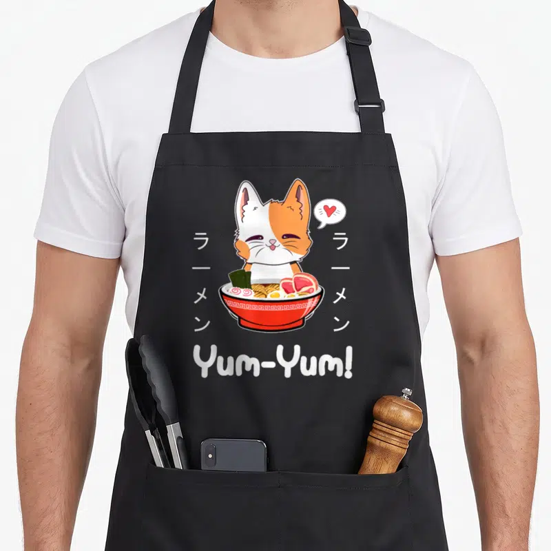 Tablier de cuisine noir avec un chat bicolore mangeant du ramen et le texte Yum-Yum en blanc.
