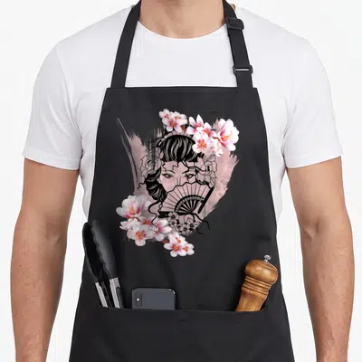 Tablier de cuisine noir avec illustration d'une Geisha japonaise tenant un éventail, encadrée par des fleurs de cerisier roses et blanches.