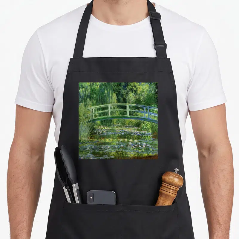 Tablier de cuisine noir avec un pont vert enjambant un lac de nénuphars blancs et roses.