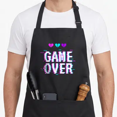 Tablier de cuisine noir avec le texte GAME OVER en pixel art blanc et des cœurs brisés roses et bleus au-dessus.