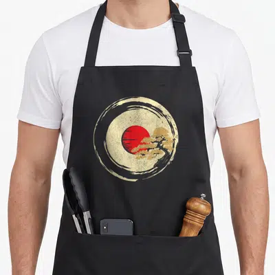 Tablier de cuisine noir avec illustration circulaire d'un soleil rouge et d'un bonsaï doré sur fond texturé beige.