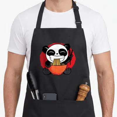 Tablier de cuisine noir avec un panda anime mangeant des nouilles ramen dans un bol orange sur fond rouge.