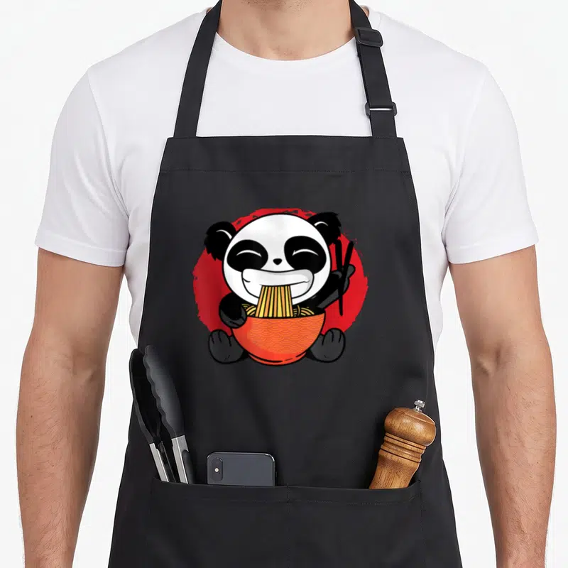 Tablier de cuisine noir avec un panda anime mangeant des nouilles ramen dans un bol orange sur fond rouge.