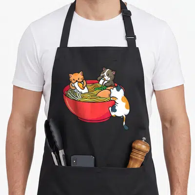 Tablier de cuisine noir avec illustration de trois chats mangeant des nouilles ramen dans un grand bol rouge.