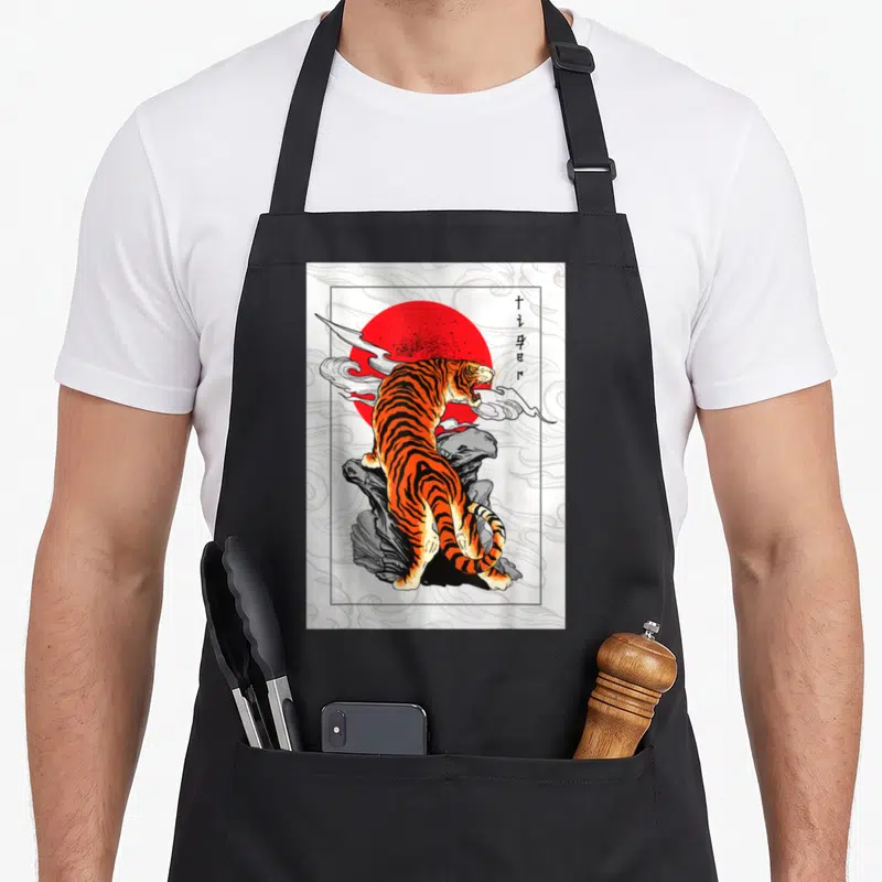 Tablier de cuisine noir avec illustration d'un tigre sur roche devant un soleil rouge et des symboles calligraphiques.