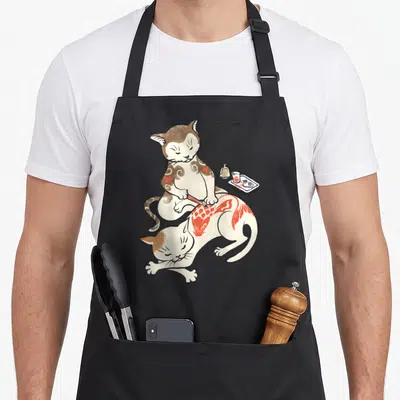 Tablier de cuisine noir avec illustration d'un chat tatoueur appliquant un motif de carpe rouge sur un autre félin allongé.