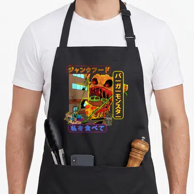 Tablier de cuisine noir avec illustration d'un monstre burger attaquant une ville et des textes en japonais colorés.