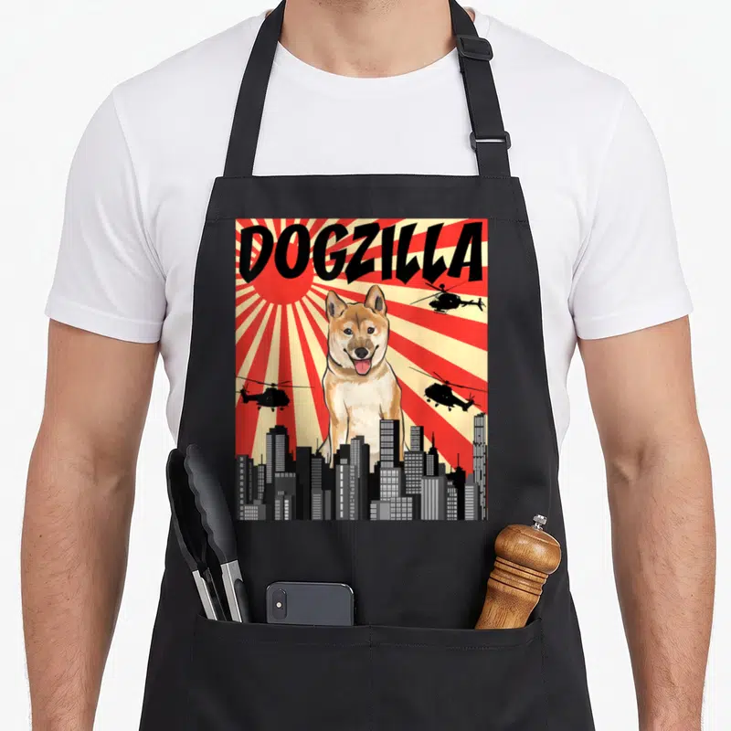 Tablier de cuisine noir avec illustration colorée d'un Shiba Inu géant nommé Dogzilla sur fond de soleil rayonnant et hélicoptères.