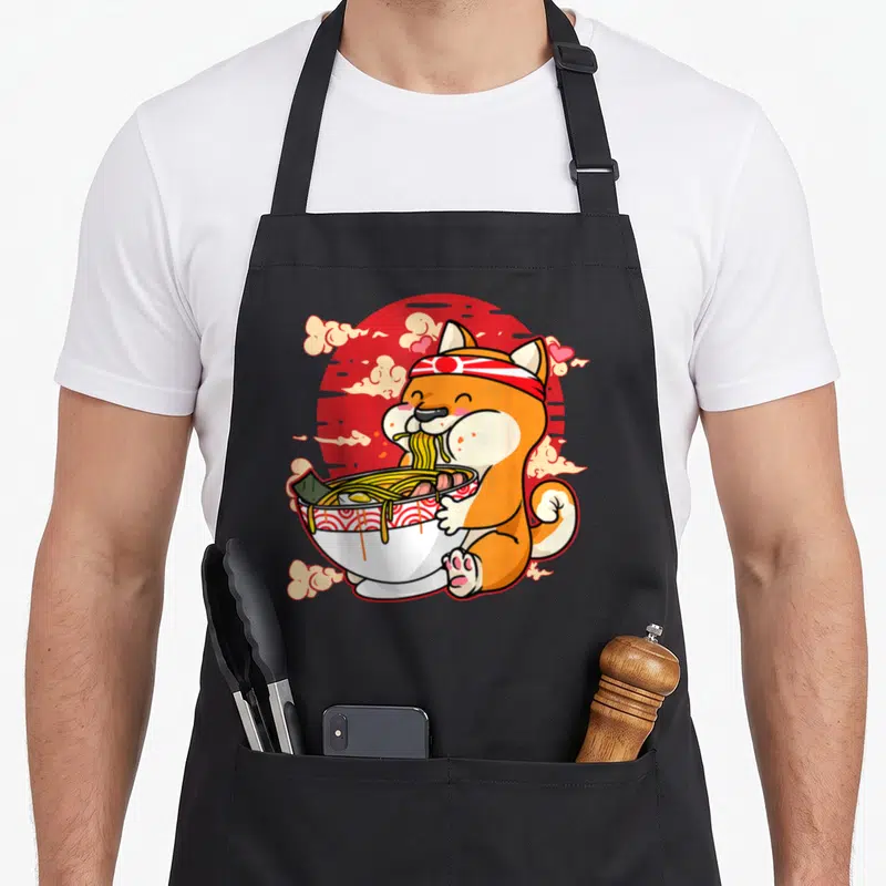 Tablier de cuisine noir avec illustration d'un chien Shiba Inu mangeant des nouilles de ramen devant un soleil rouge.