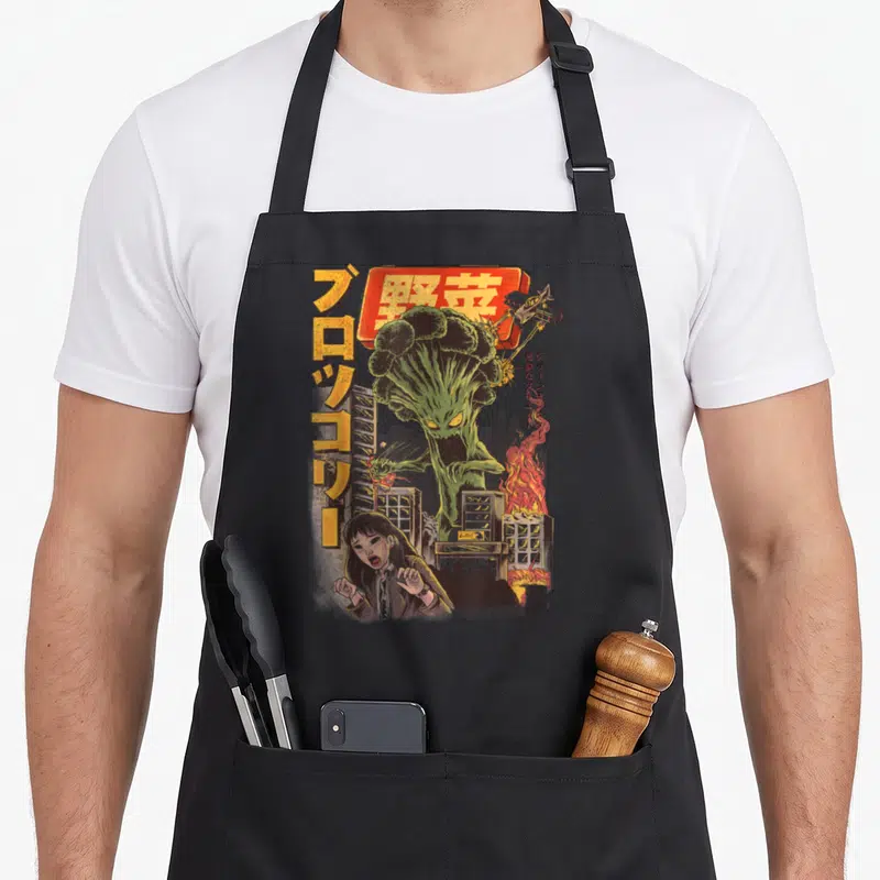 Tablier de cuisine noir avec illustration d'un monstre brocoli détruisant une ville japonaise et une femme effarée.
