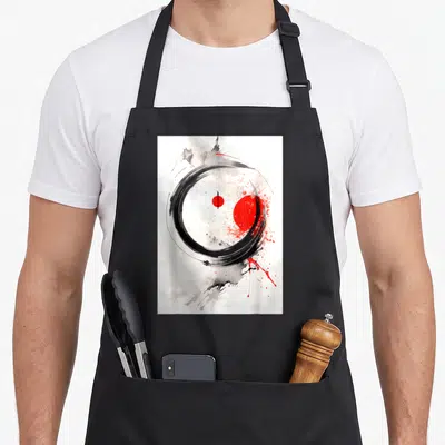 Tablier de cuisine noir avec un grand cercle Enso noir, une sphère rouge et des éclaboussures artistiques sur fond blanc.