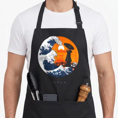 Tablier de cuisine noir avec une illustration circulaire montrant une vague, une geisha et un temple japonais sur fond orange.