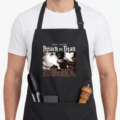 Tablier de cuisine noir avec logo Attack on Titan, illustration de titan et caractères japonais en bas.