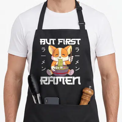Tablier de cuisine noir avec un Shiba Inu mangeant du ramen et le texte But First Ramen en blanc.