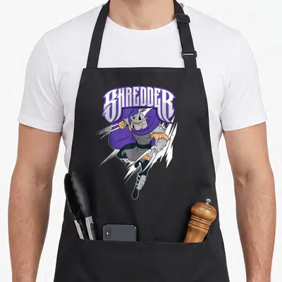 Tablier de cuisine noir avec illustration colorée de Shredder en mouvement dynamique et texte stylisé au-dessus.