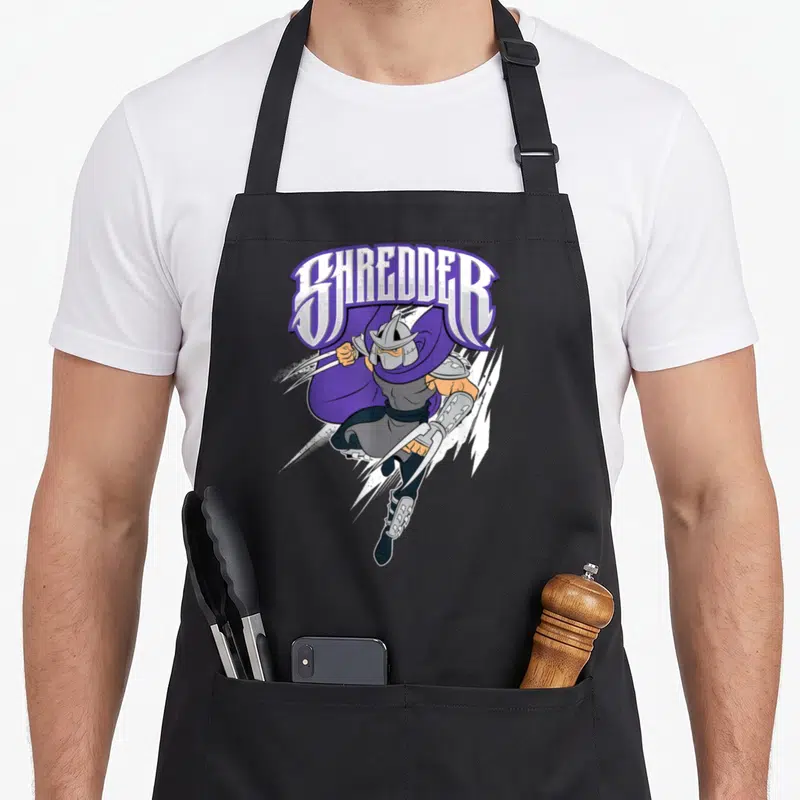 Tablier de cuisine noir avec illustration colorée de Shredder en mouvement dynamique et texte stylisé au-dessus.