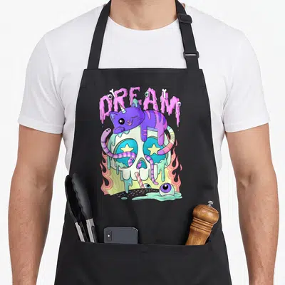 Tablier de cuisine noir avec illustration d'un caticorn violet sur un crâne aux yeux en étoiles et texte rose coulant.