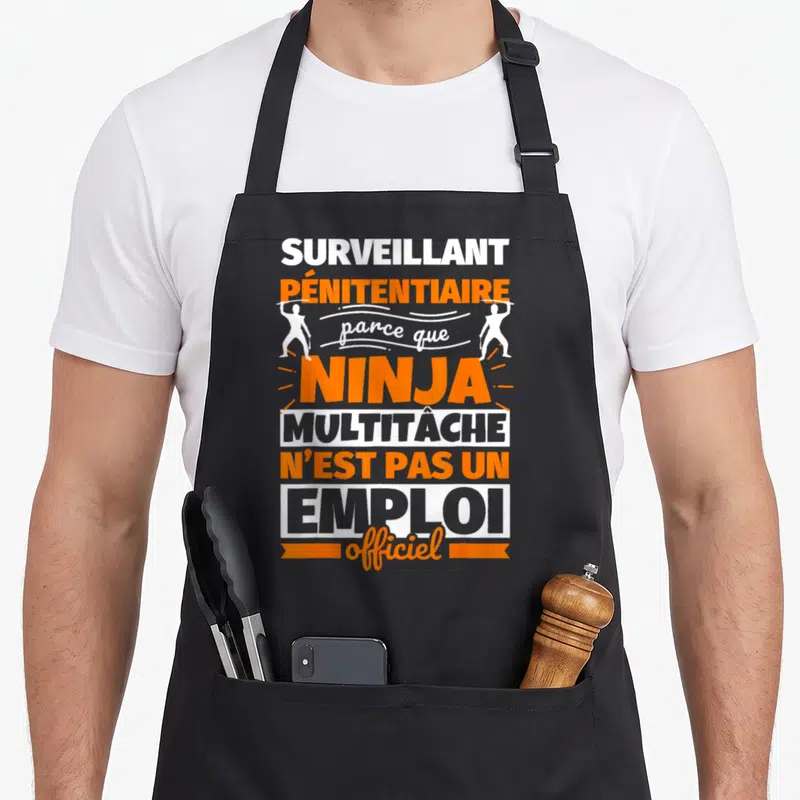 Tablier de cuisine noir avec le texte Surveillant pénitentiaire parce que ninja multitâche n'est pas un métier reconnu en orange et blanc.