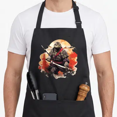 Tablier de cuisine noir avec illustration d'une tortue samouraï ninjatenant une katana devant un soleil orange.