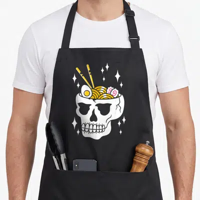 Tablier de cuisine noir avec illustration d'un crâne blanc rempli de nouilles jaunes, d'un œuf et de baguettes dorées.