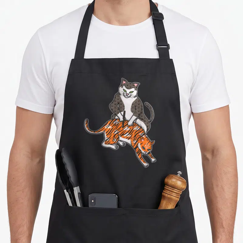 Tablier de cuisine noir avec illustration d'un chat artiste tatouant un autre chat en tigre orange.
