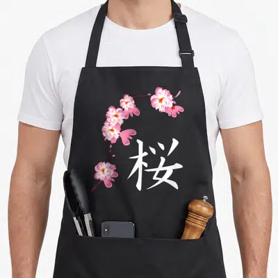 Tablier de cuisine noir avec une branche de fleurs de cerisier roses et le kanji blanc Sakura imprimé au centre.