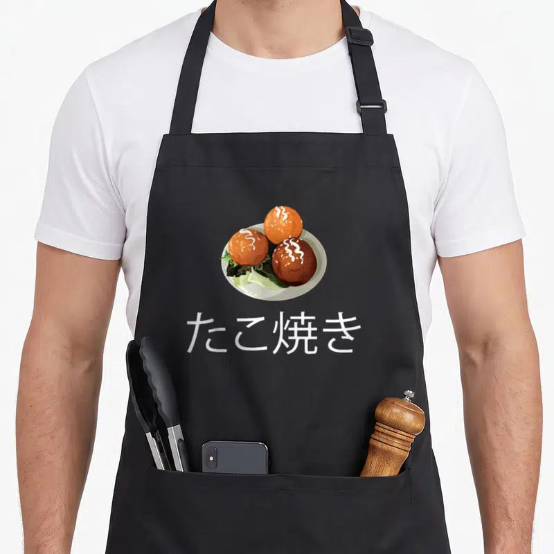 Tablier de cuisine noir avec illustration de trois takoyaki sur assiette et texte en kanjis blancs.