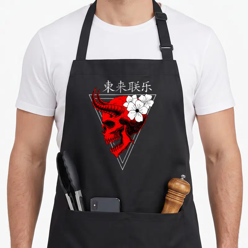 Tablier de cuisine noir avec tête de démon rouge, fleurs blanches et triangles blancs superposés.